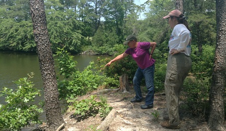 Two people standing on a wooded shoreline, one pointing toward the water.