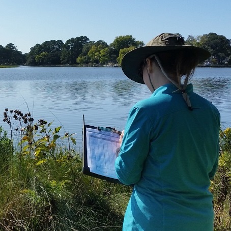 Person wearing a wide-brim hat recording data on a clipboard while standing by a shoreline with water and vegetation.