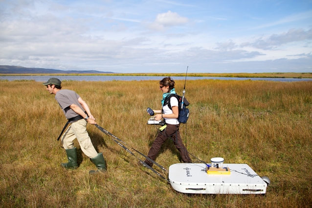 Two field scientists using ground penetrating radar system over grassy marshland; woman in front is dragging the antenna across the ground transect, and another woman walks alongside while operating the controller.