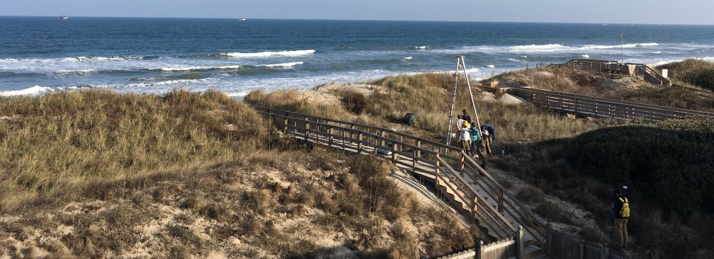 Field scientists using vibracore tripod system to collect sediment cores on a sweeping dune ridge, with ocean waves crashing in the background.