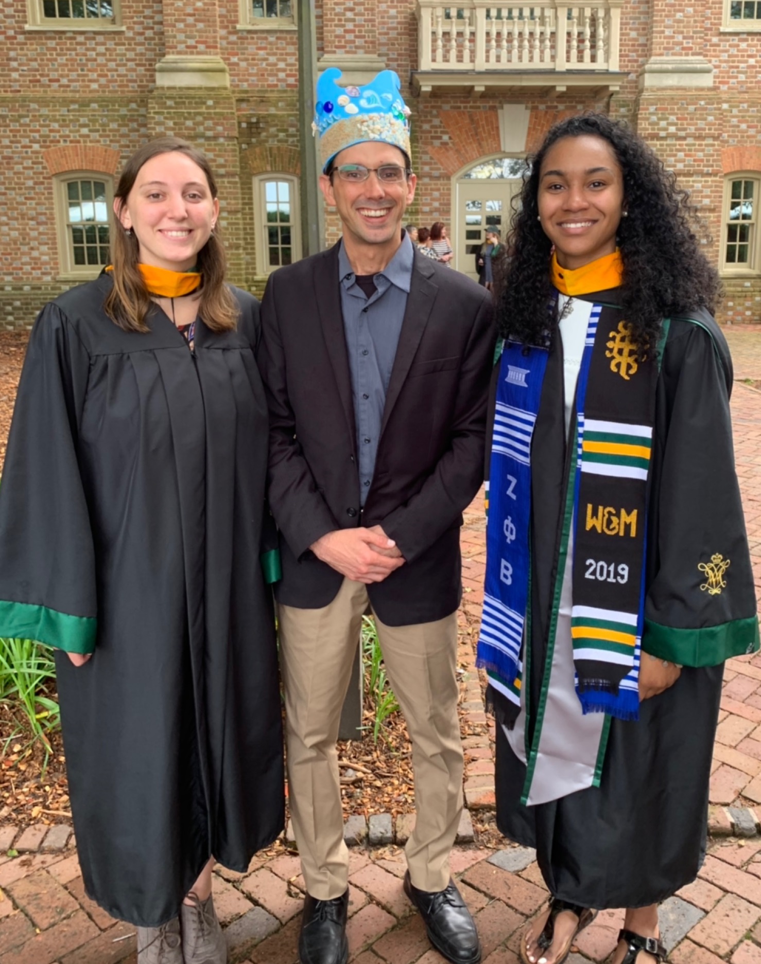 Chris hein poses with two graduating students while wearing a jaunty crown hat