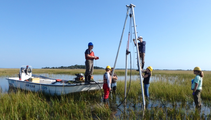 Group of field scientists stand around tripod vibracore rig during sample collection in flooded coastal marshland, with skiff boat used for transport in the background.